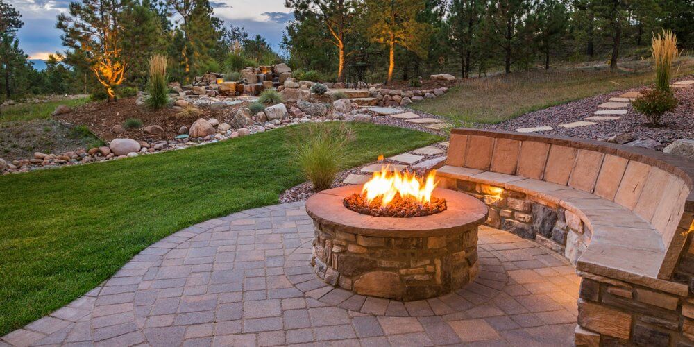 A stone fire pit with built-in bench seating on a paved patio, with a landscaped yard and trees in the evening light.