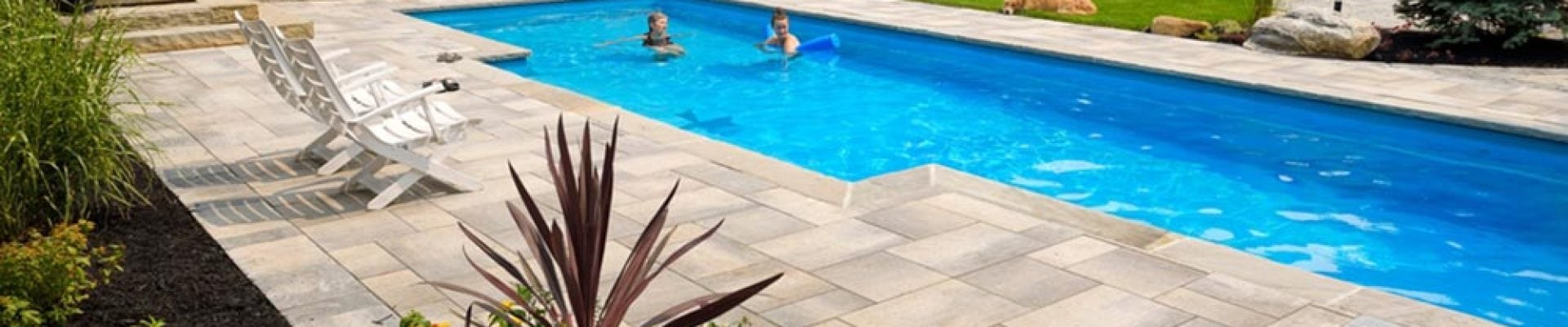 A landscaped backyard features a rectangular swimming pool with two people swimming, lounge chairs, potted plants, and a wooden fence in the background.