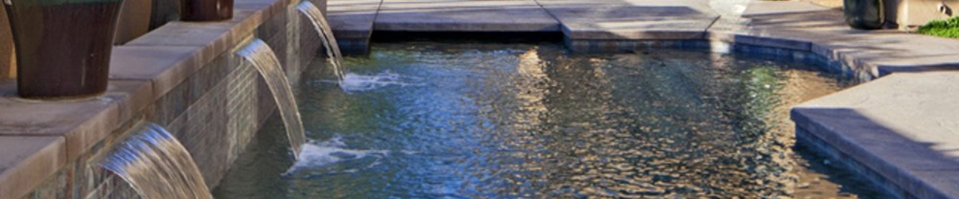 A luxurious backyard swimming pool with waterfall features, surrounded by potted plants. There is a hot tub in the background, shaded by an umbrella.