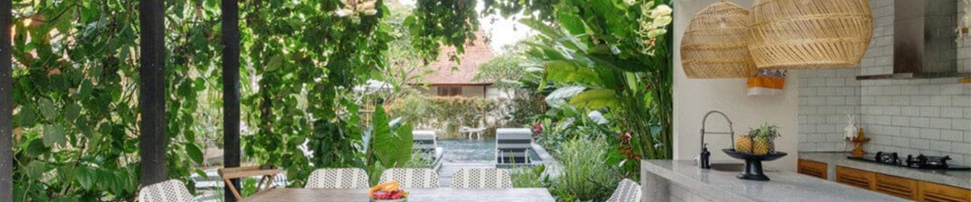 Outdoor dining area with a table and eight chairs under a glass roof, surrounded by greenery. A nearby kitchen counter features hanging woven lamps and a tiled backsplash.