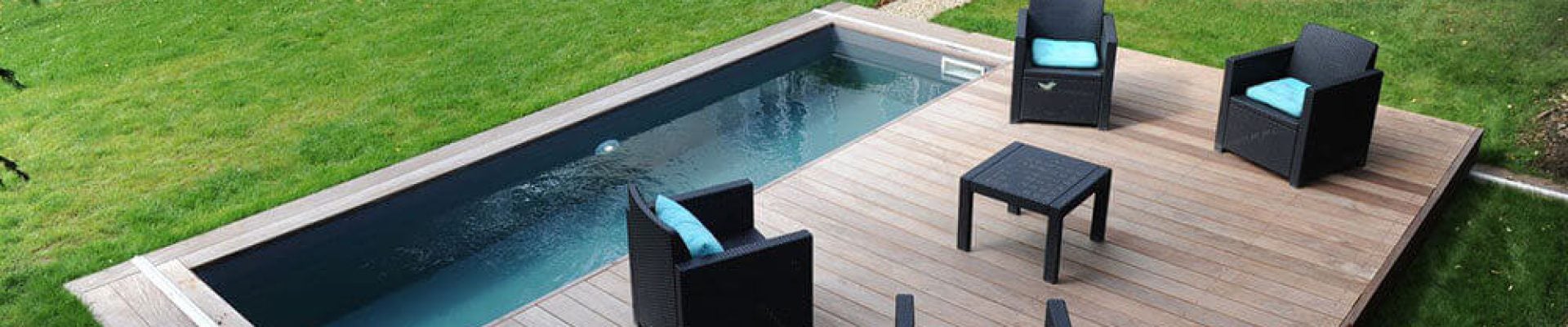 Backyard with a rectangular pool and adjacent wooden deck featuring four black chairs with blue cushions and a black table. Grass surrounds the deck, with two green chairs in the background.