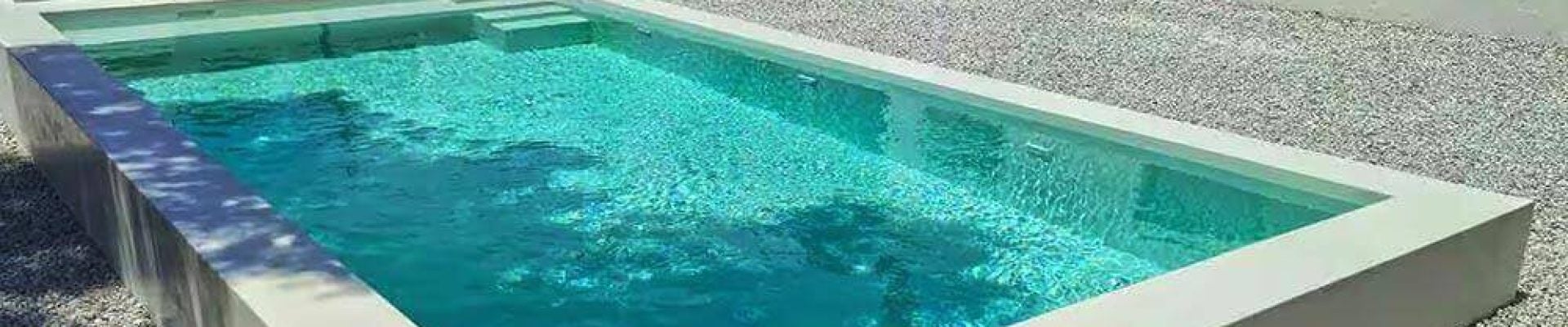 A rectangular in-ground swimming pool in a modern, minimalist backyard with white walls, gravel ground cover, and sparse greenery.