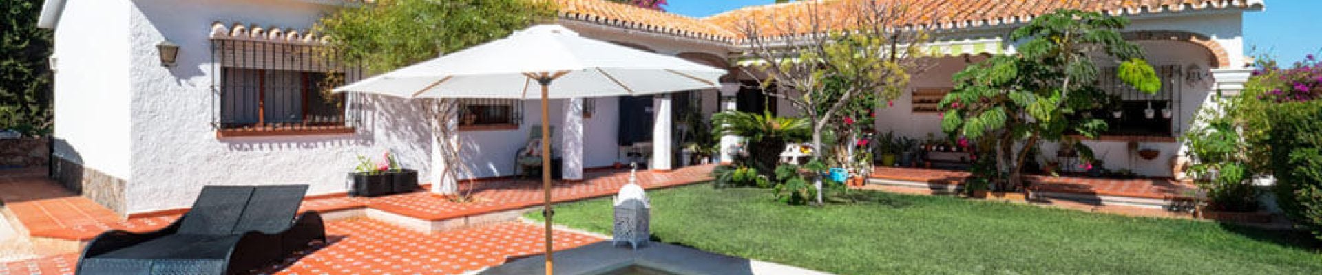 A white villa with a terracotta-tiled roof features a pool with a large umbrella, sun loungers, and a garden area under a clear blue sky.