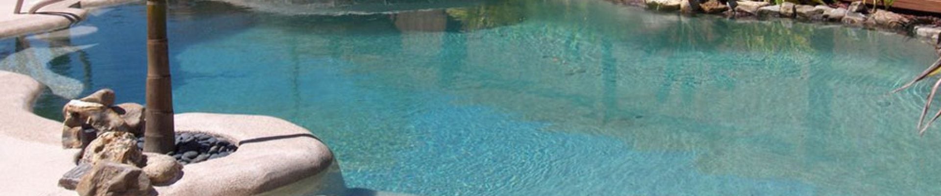 A backyard swimming pool with clear water, surrounded by a wooden fence and landscaped with low shrubs and plants. Two lounge chairs sit on a stone deck nearby.