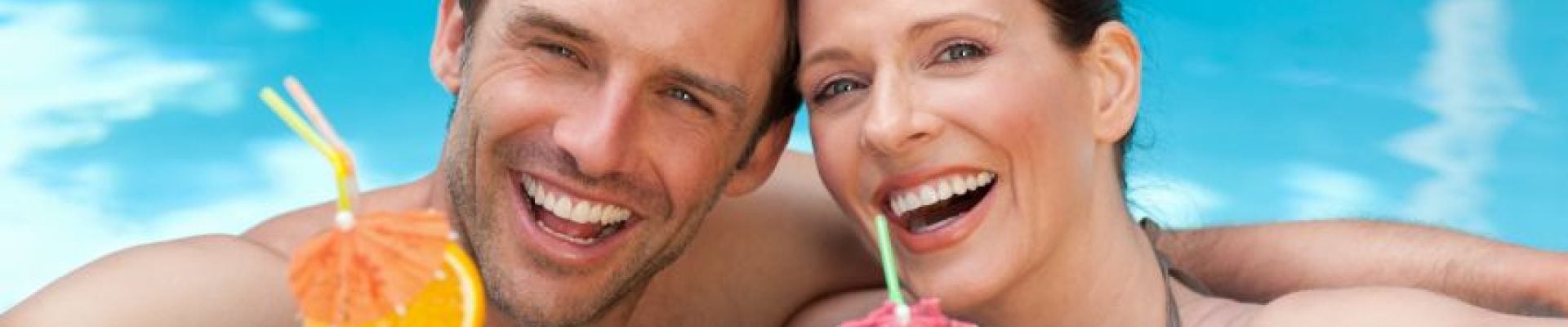 Two people smiling while leaning on the edge of a pool, holding colorful drinks with umbrellas and straws.