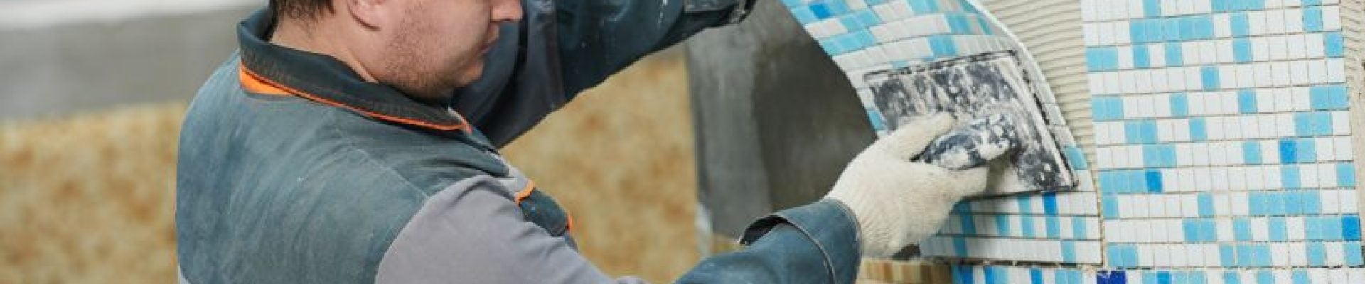 A construction worker wearing a white hard hat and gloves applies tiles to a wall using a trowel.
