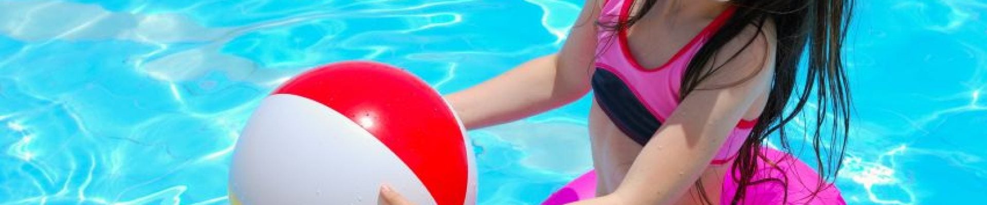 A young girl in a pink and black swimsuit and pink swim ring holds a colorful beach ball while standing in a swimming pool, smiling.