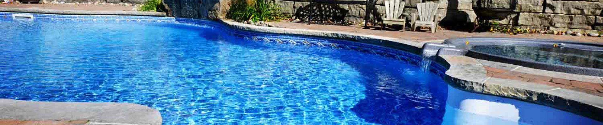 A backyard pool with clear blue water, stone steps leading into the pool, a small waterfall feature, and surrounding landscaping. A hot tub is visible to the right. A fence encloses the area.