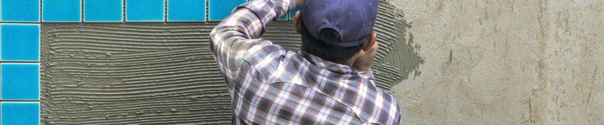 Person in a plaid shirt and blue cap applying blue tiles to a wall using mortar.