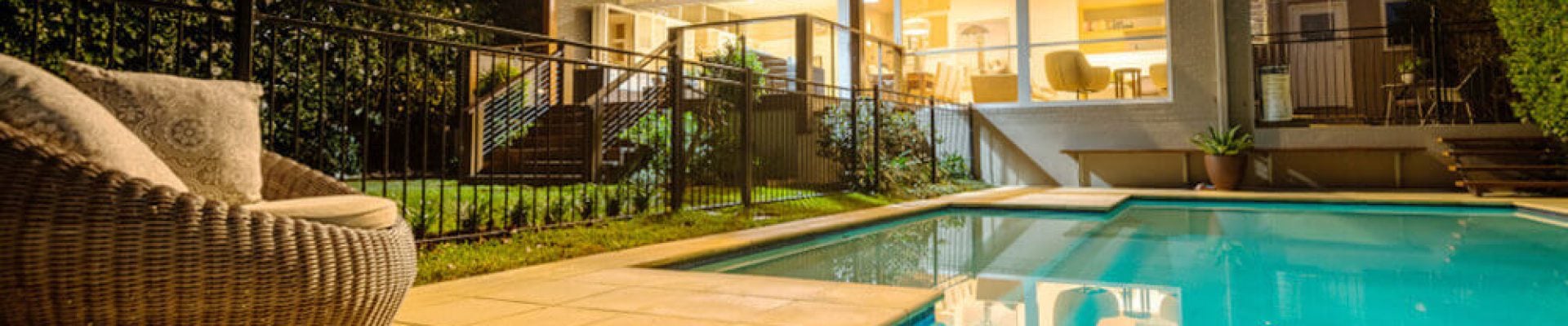 A modern two-story house with large windows is illuminated at night. It features an outdoor swimming pool surrounded by a patio and foliage.