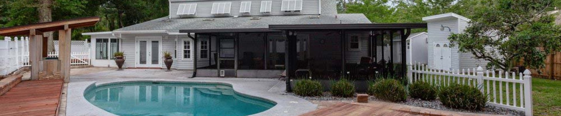 A backyard features a kidney-shaped pool with a surrounding wooden deck, a screened porch area, and a white house in the background under a clear sky.